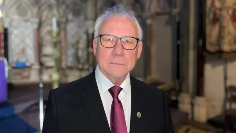 BBC Mr Lloyd-Jones looks into the camera. He is not smiling. He wears black rim glasses, a black suit jacket and a red tie. Behind him the church walls can be seen. 