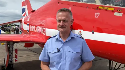 Sqn Ldr Dave Kerrison is pictured in front of one of the Red Arrow planes wearing a light blue shirt with military patches on his shoulders. He is clean shaven with short grey hair.