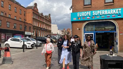 Laura Coffey/BBC A police officer with three women walking down a high street. The police officer is pointing a talking to one of the women. There are other officers in the background and a shop called "Europe Supermarket".