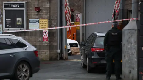 PA Media A still of Lurgan police station with a the front of a white Audi car parked inside the station. There is a brown piece of paper covering the license plate. There is a red cordon closing off the station. In the distance is a police officer standing with their back turned to the camera.
