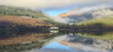 Tim Berrall A grand house on the edge of a loch, nestled in trees with hillsides around it, reflected on the still water, under a cloudy blue sky.