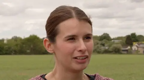 Shaun Whitmore/BBC Adriana Hudson looks away from the camera while standing outside near a field. She has long dark hair that is tied back behind her head. She wears a red knitted jumper and black top underneath.