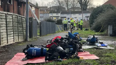 Breathing apparatus used for firefighting lies on the ground outside the property, which can be seen in the background on the left behind a tall wooden fence. Much of the red brickwork that can be seen is blackened.  There are a number of fire officers outside the house in yellow jackets and helmets.