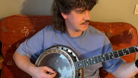 Stephen McKee is sitting on a red sofa with an abstract cream and green pattern. He has a moustache and dark curly hair which comes down over his forehead and is also long in the back. He is wearing a light blue t-shirt and is playing a banjo. He is looking up towards his left hand which is holding the strings, while he plucks with his right hand. 