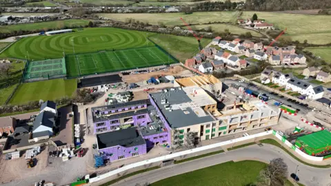 An aerial view of a school being constructed. There is purple panelling on one building and a crane lifting panelling on another. The school is surrounded by a new housing development and sports pitches.