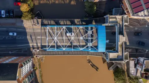 The bridge has a blue metal criss-cross casing over it. The brown water of the River Hull runs beneath the bridge.