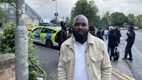 A man in a white polo shirt and beige jacket stands on a pavement. There are police cars in the background.