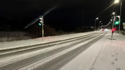 BBC Snow on the road. There are traffic lights and cars in the distance.