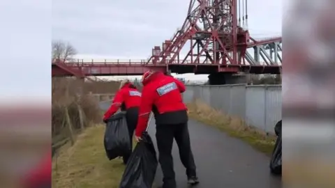 A man and a woman picking up litter and putting it into black bags while walking along a riverside path, with Middlesbrough's Newport Bridge in front of them.