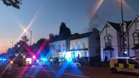 Blue and pink lights of emergency services vehicles are flashing, with police cars and fire engines outside a row of white buildings