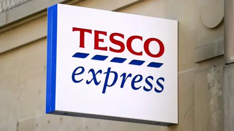 A white rectangular sign that reads: Tesco Express. The Tesco word is red, there are five blue dashes underneath each letter and the word express is in blue underneath that. It is hung on a beige brick wall. 