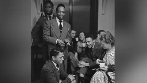United States Air Force personnel attend an officers' club dance at RAF Lakenheath. It is a black and white photo. The men are in suits and ties, and a woman is in the foreground in a dress, with curly hair and holding a mug.