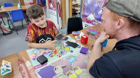 A young boy plays Pokemon cards with a fellow competitor. They are sat at a table with some of the cards laid out in front of them on colourful mats.