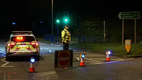 Police at the scene of a road traffic accident in Robertsbridge, East Sussex. One police officer is stood in front of a police car which is advising that the road is closed. There are a number of cones on the road.