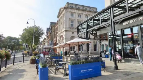 Stand Up for Southport A view of Lord Street, Southport in summer with tables and chairs with beige umbrellas outside as bistro. There are people walking along the road and a person with a baby in a pram looking in a window in the background.