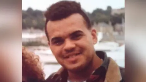 Archive photograph of Tyrone France, who smiles at the camera. He has brown curly hair, brown eyes and stands on a bridge outside. He wears a blue t-shirt, red shirt and a coat. 