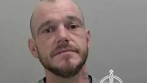 Mugshot of Luke Simon wearing a grey jumper. He has short hair and a stubble
