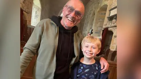 Film director Danny Boyle wearing large framed glasses and a black hoodie in a church with his arm around a young blonde boy wearing a blue dressing gown and a pyjama top with space craft on it.