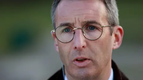 PA Media Andrew Muir, a man with short grey hair and blue eyes, speaks to the media. He is wearing large, round metal-framed glasses and a brown jacket over a pale shirt. The background of the close-up photo is blurred.