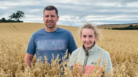 Watson family Fergal Watson with his teenage daughter standing in a large crop field on a cloudy day. He has short, fair hair and is wearing a blue t-shirt. She has curly blonde hair, tied back and is wearing a beige and peach jacket. 