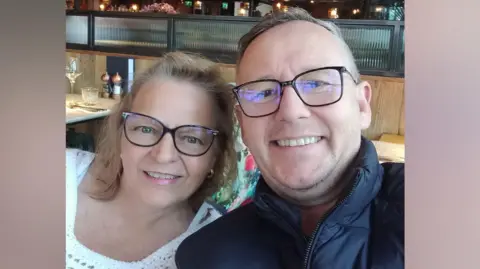 Alan Bruce Denise and Alan Bruce. Denise wears a white crocheted top, whilst Alan wears a navy blue zip jacket. Both have dark-rimmed glasses and are both smiling at the camera
