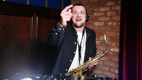 A man points at the camera while wearing a white tshirt and dark denim overshirt. He is wearing glasses and headphones and has short dark hair. He is holding a saxophone in one hand and pointing with the other.