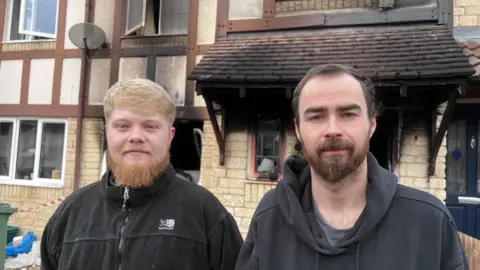 Liam and Harry stand in front of the burned remains of their home. the doors and windows are missing and there are black smoke stains up the yellow stone walls. They have beards and are wearing black jumpers