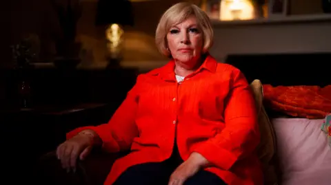 STV Studios Factual/BBC A woman wearing a bright red, button-up shirt, seated indoors on a sofa with a cushion and throw blanket, in a warmly lit living room setting