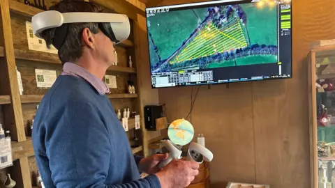 Ian Sexton is standing in a shed wearing virtual reality goggles while he looks at a screen which shows fields. One of the fields has a yellow grid in laid over it. In his hands are two handsets.