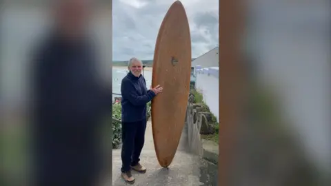 An elderly man with grey hair and a beard holding up a coppery toned fibre glass surf board and smiling at the camera with the beach behind him and grey sky