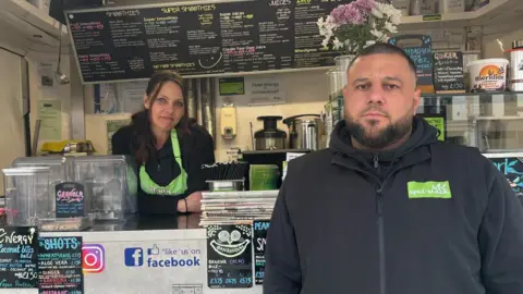 BBC A woman and a man at a food truck. There are menu boards behind them and they are both wearing black and green uniforms. 