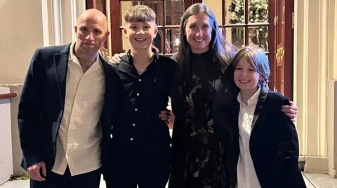 Family photograph Ben, far left, has a shaved head, and is wearing a dark jacket and white shirt. His son Charlie, second left, has light brown and is wearing a black shirt. Ben's wife is wearing a black dress and has long brown hair. The couple's other son, Dexter, is on the far right of the picture and is wearing a black jacket and white shirt.