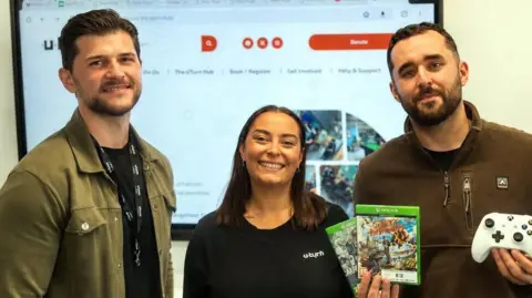 Two men stand either side of a woman. All three are smiling at the camera. The man on the left is wearing a green jacket, black top and a black lanyard. The woman in the middle is wearing a black top with a white logo that says UTurn. The man on the right is wearing a green top and is holding two video games and a console controller. 