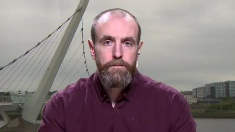 Mark Durkan - a man with receding hairline and a brown beard streaked with grey. He is wearing a plum coloured shirt and is sitting against a background of the peace bridge in Londonderry.