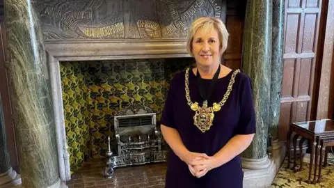 LDRS A woman with short blonde hair is wearing a navy blue dress and large gold ornate mayoral chains around her neck. She is standing in front of a large Victorian stone fireplace in a room in Sheffield Town Hall.