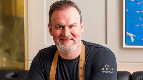 Glynn Purnell - a man with short dark hair and a grey beard - smiles as he sits on a leather chair wearing a black shirt with a brown apron.