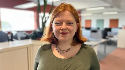 Rachel Hart is smiling at the camera. She has ginger hair in a long bob and is wearing a khaki top and multi-coloured beads. She has a nose ring