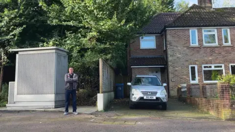 Brett Kemp, a man in his 50s, standing next to a tall grey shed that houses a toilet, erected for the convenience of bus drivers. His arms are folded and he looks cross.