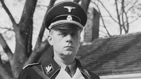 Getty Images A black and white photo of a man in dark military uniform with a dark cap with a metal bird badge on it and the roof of a house behind him