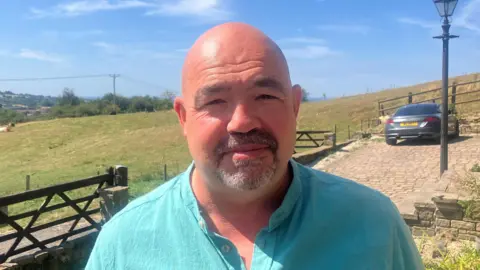Alan Hopwood is wearing a blue/green shirt. He has a bald head and a goatee beard. In the background is a field and a driveway.