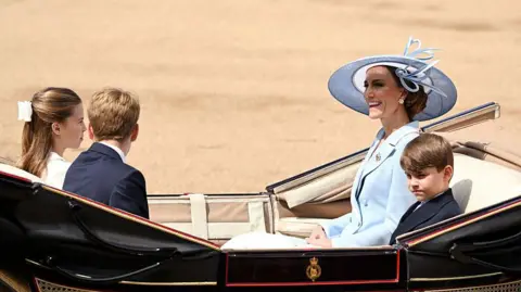 Getty Images Princess Catherine sits in a carriage with a tan interior with Prince Louis, who sits next to her, and Princess Charlotte and Prince George, who sit opposite her. She is smiling and wearing a pale blue jacket with a pale blue hat. Louis and George are wearing dark suits and Charlotte is in white, with a white bow in her hair.