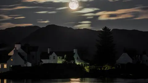 Sam Bilner Full moon shines over white cottages and mountains, reflecting on a still lake.