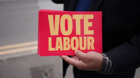 A close up picture of a persons arm holding a pile of vote labour flyers