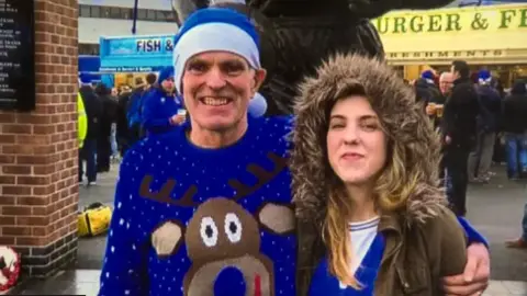 A man and woman stand outside a football ground on a wet day. The man, Noel McNally, is wearing a blue Christmas jumper with a large reindeer motif and a blue and white hat. The woman, his daughter Natalie McNally, is wearing a blue and white football jersey under a brown overcoat with a brown fur hood. Her father's hand is placed around her shoulder.