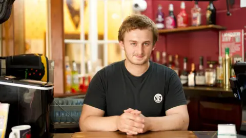 BBC Alex Barry, a young man with blond hair, stands behind an old fashioned bar in a pub.