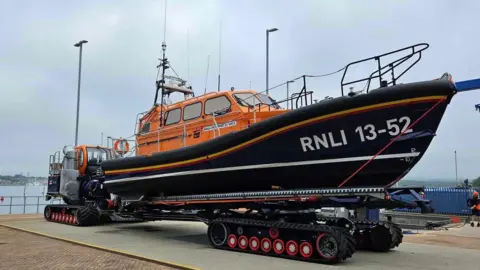 RNLI/Mark Walsham A lifeboat, which has an orange cabin and a navy hull with the lettering "RNLI 13-52" on it. The boat is on a platform made of wheels at a dockyard, with the sea behind it. It is a grey, cloudy day.