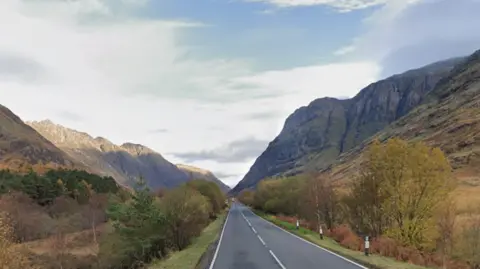 The A82 running through the Great Glen, with mountains on either side of the flat road, and a blue sky with light cloud above.