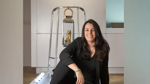 Simon Jacobs/St Albans Museum Maria Meyer sits in front of one of her exhibits - a Zimmer frame with a lantern hanging from the top, with a small sculpted dog inside it. Behind the exhibit is a cream wall with panels and a wooden floor.