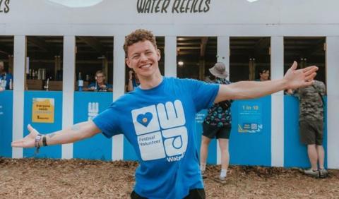 Conor Blundell wearing a blue wateraid t-shirt, standing with his arms outstretched and smiling in front of a water refills station at Glastonbury