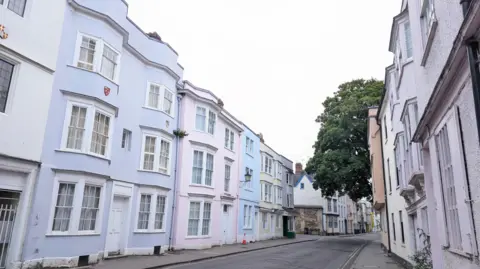 Lucie Johnson MONDAY - A street in Oxford with pastel coloured houses on each side with large traditional white windows. A large green tree overhangs the street half way down and overhead the sky is white.